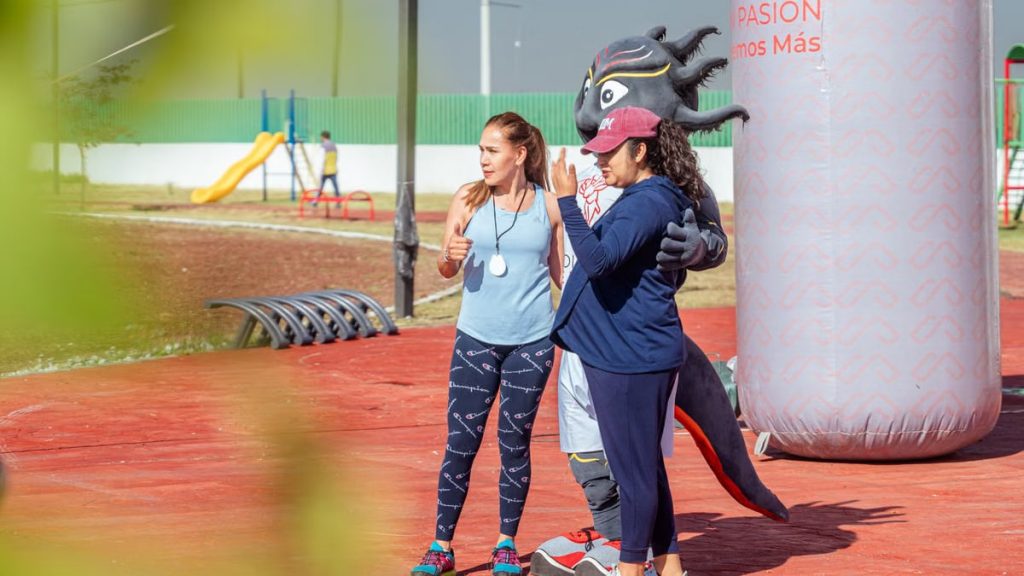 Women posing with a mascot at an outdoor sports event on a sunny day.